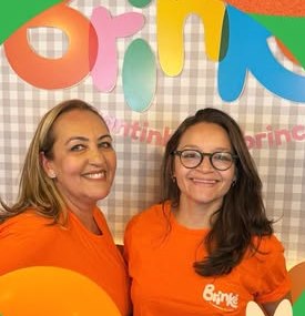 Simone e Karol, de camiseta laranja do Brinkê, em frente ao letreiro colorido